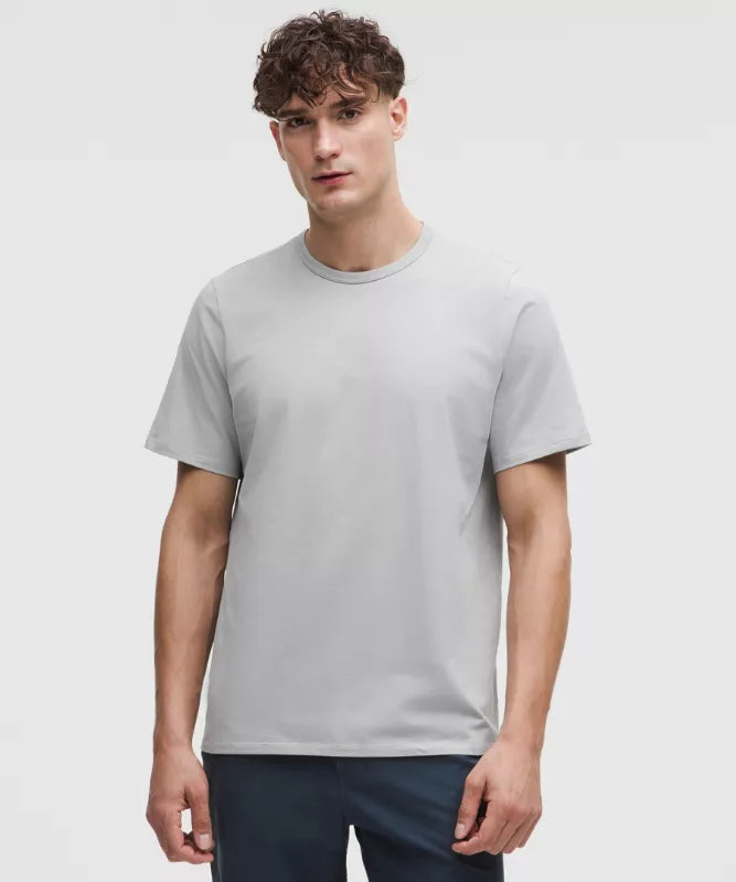 A man in a light gray t-shirt stands confidently, wearing casual navy pants against a plain backdrop.