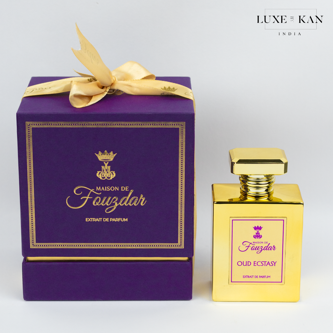 A luxurious purple box adorned with a gold ribbon contains a golden perfume bottle labeled "Oud Ecstasy" by Maison de Fouzdar.