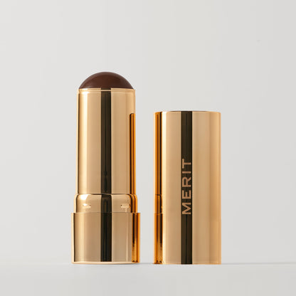 Merit beauty bronze balm sheer bronzer makeup stick with a rounded chocolate-colored tip, partially out of its matching case.