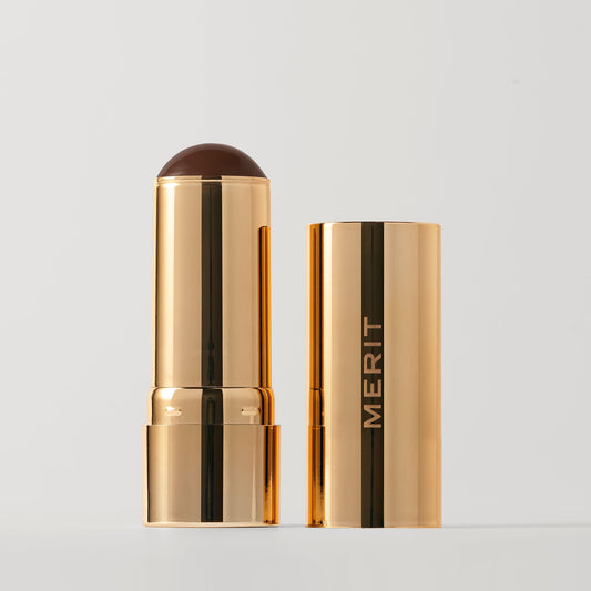 Merit beauty bronze balm sheer bronzer makeup stick with a rounded chocolate-colored tip, partially out of its matching case.
