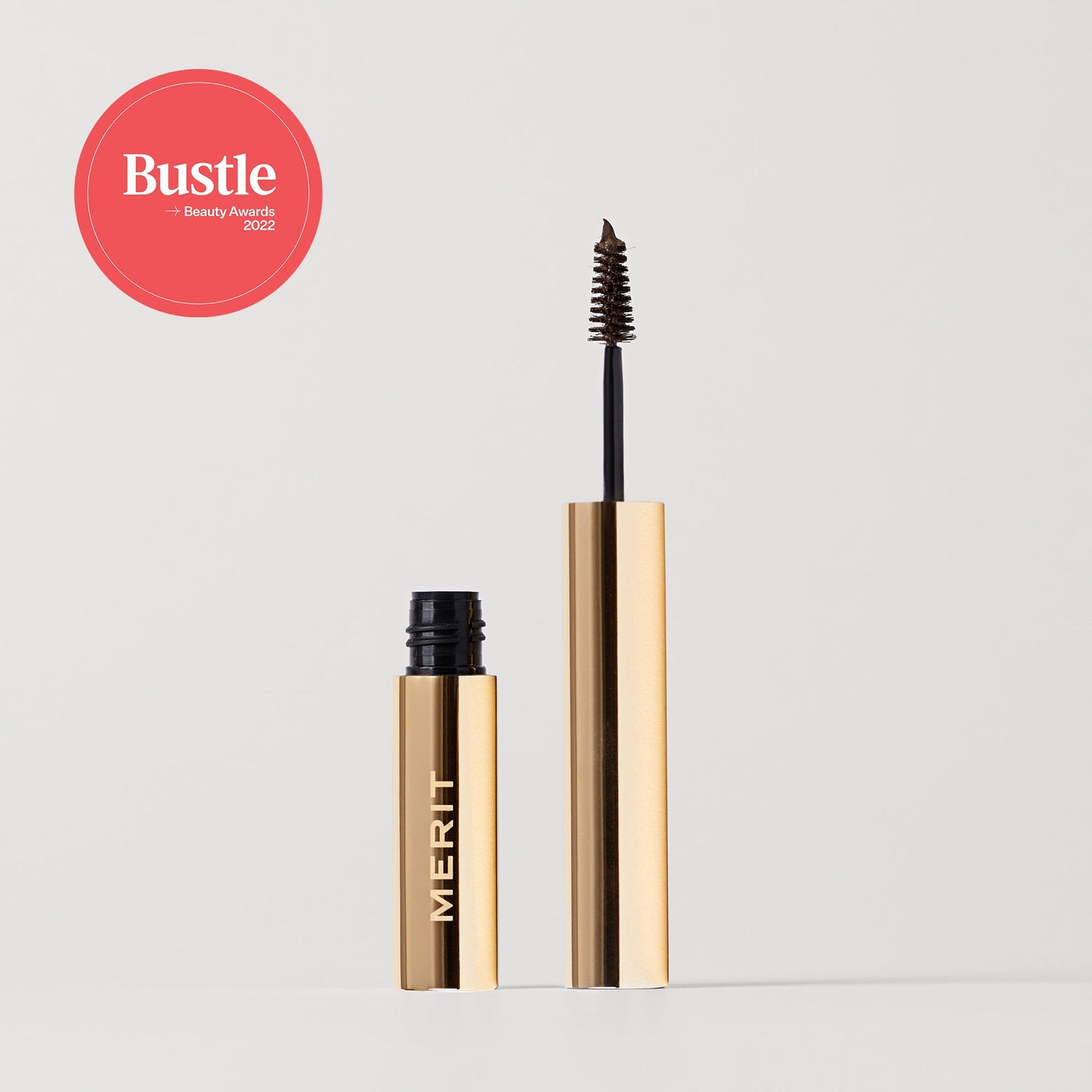 Merit beauty brow 1980 volumizing pomade with a spoolie brush, beside its sleek tube, featuring the Bustle Beauty Awards 2022 label.