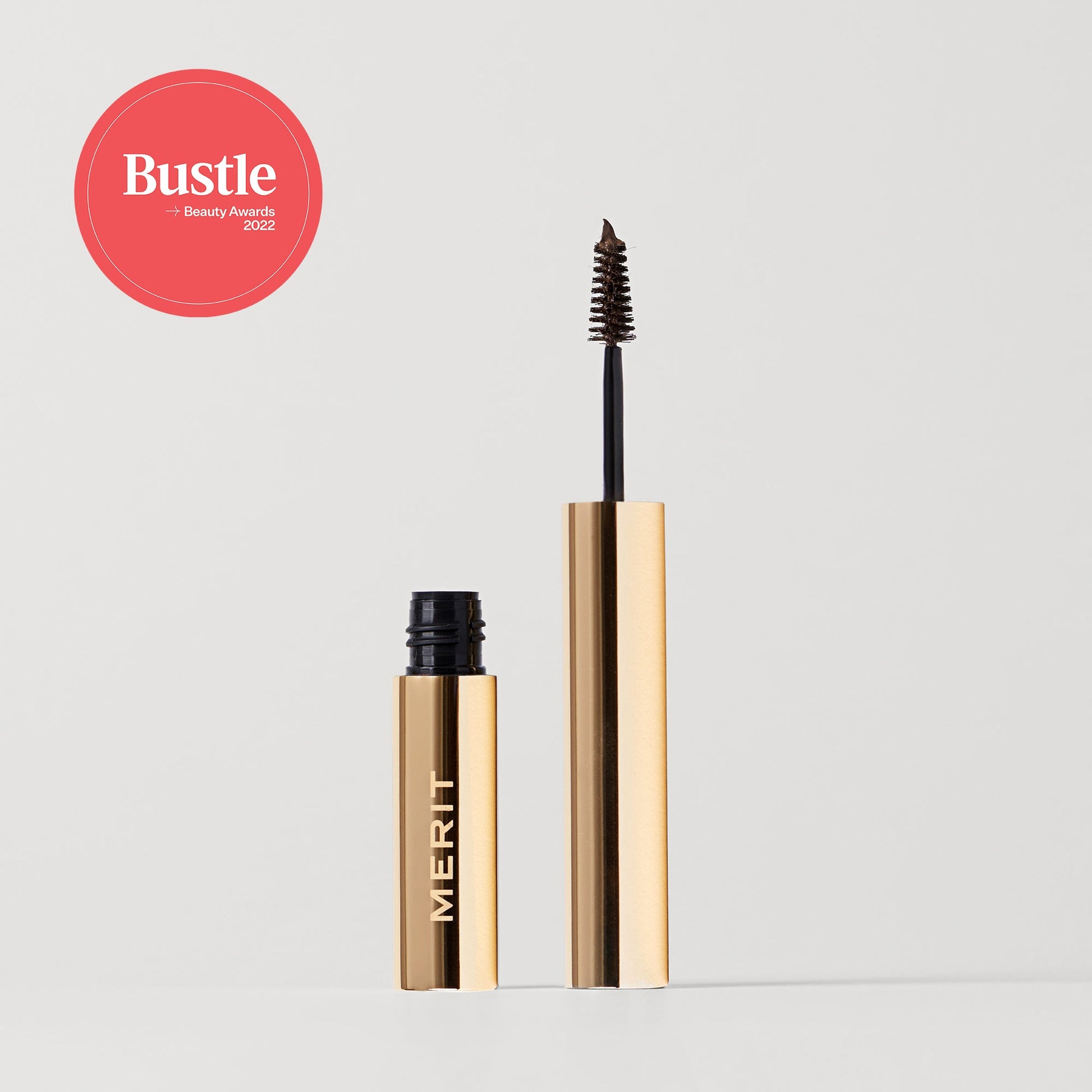 Merit beauty brow 1980 volumizing pomade with a spoolie brush, beside its sleek tube, featuring the Bustle Beauty Awards 2022 label.