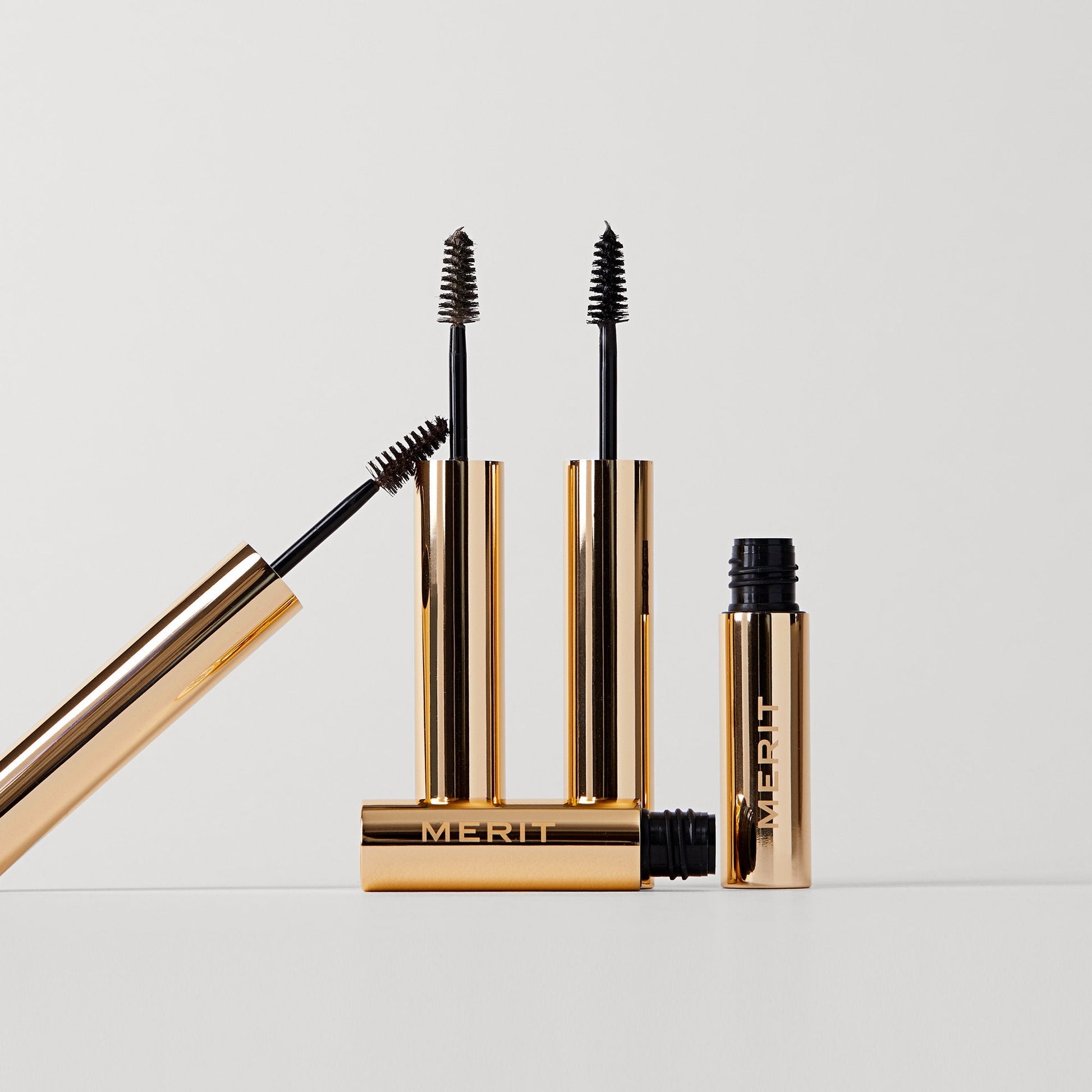 Three gold mascara tubes with black brushes, arranged artistically on a white background, showcasing MERIT branding.