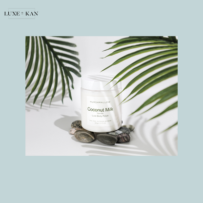 A jar of Murshmallow Coconut Milk Luxe Body Polish sits atop smooth stones, surrounded by green palm leaves on a light backdrop.