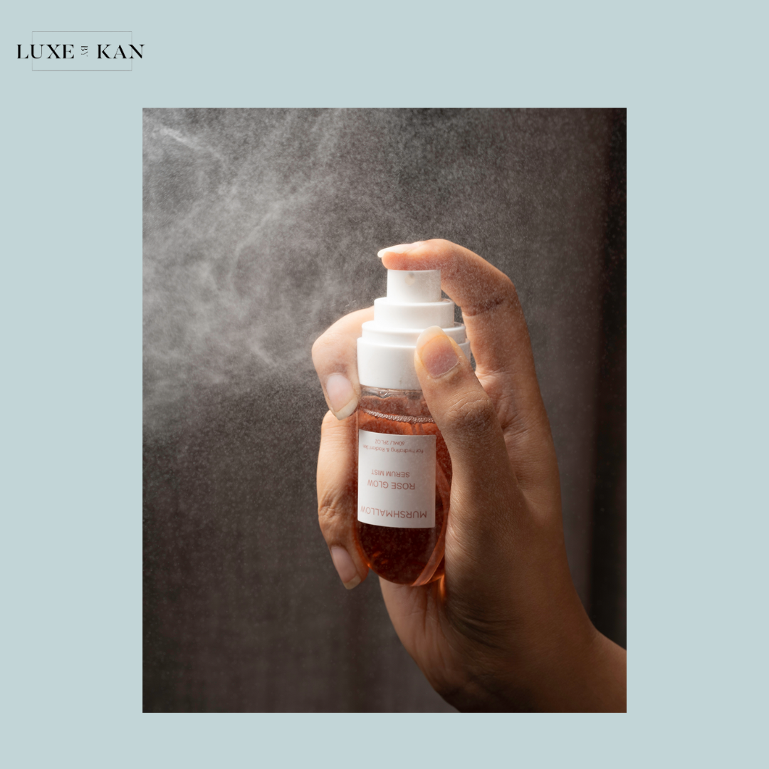 A hand holding a spray bottle releasing a fine mist of rose glow serum.