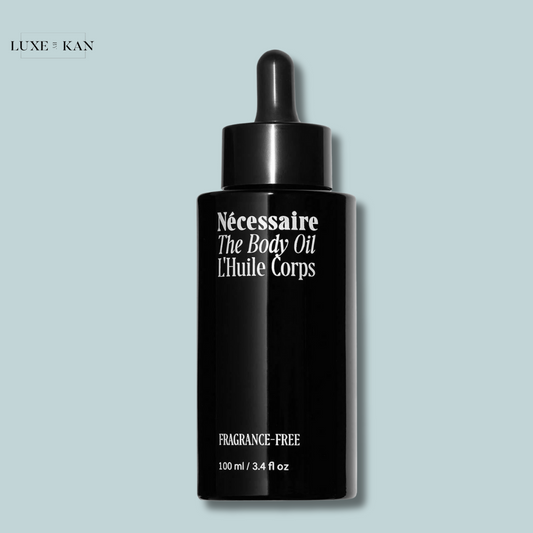 A sleek black bottle labeled "Nécessaire The Body Oil" with a dropper cap, on a soft blue background, emphasizing it's fragrance-free.