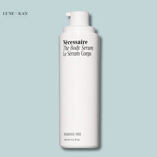 A sleek white bottle of Nécessaire The Body Serum, labeled "fragrance-free," on a soft blue background. Contains 150 ml.