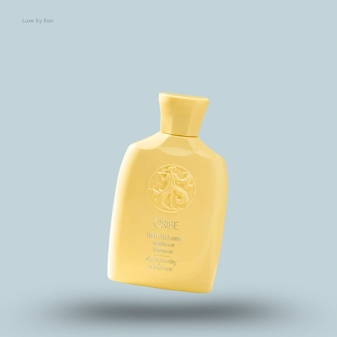 Oribe Hair Alchemy Resilience Shampoo in a yellow bottle with the brand's name and logo against a light blue background.