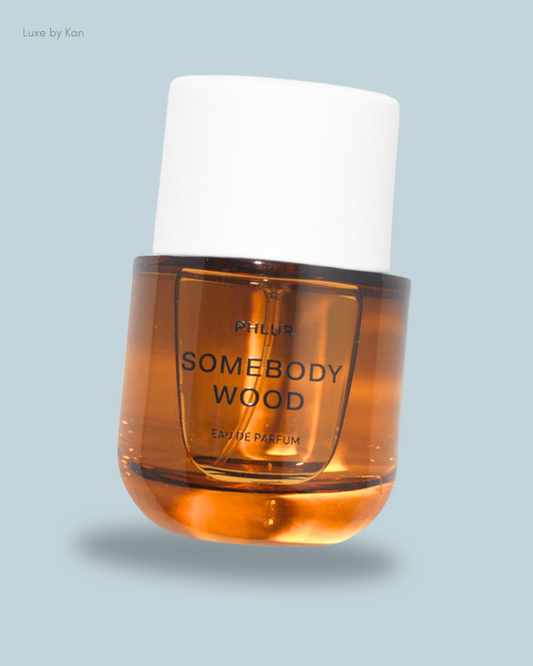 A bottle of Phlur's "Somebody Wood" eau de parfum, featuring a transparent amber liquid and a white cap against a soft gray backdrop.