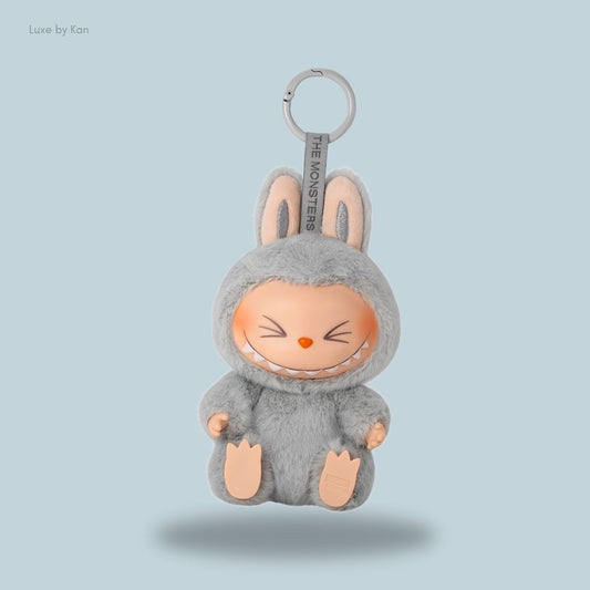 A cute gray Pop Mart Labubu Have a Seat – Hehe vinyl plush keychain doll, featuring a smiling face, bunny ears, and a tag that reads “THE MONSTERS.”