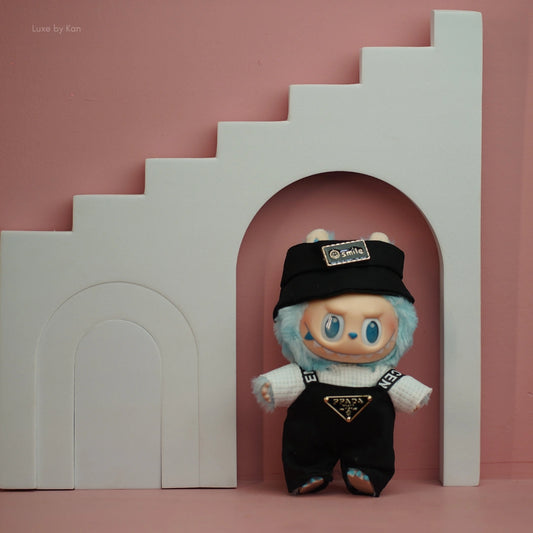 Labubu doll within a set of pink wall and white stairs props; wearing black bucket hat/Prada overalls with streetwear styled clothing.