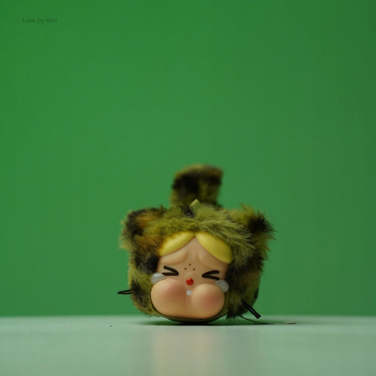 Popmart Wild Whiskers Crybaby figurine with a fuzzy green-and-brown bear-like head, blond bangs, downturned, cartoonish eyes, tears, and a concerned expression.
