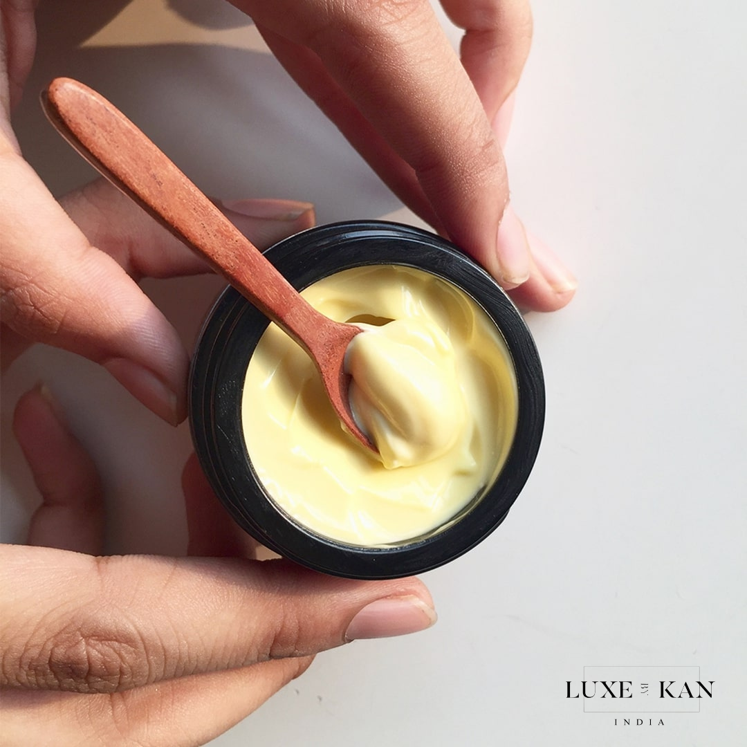 A close-up of hands holding a black jar of yellow cream with a wooden spoon, showcasing the creamy texture inside.