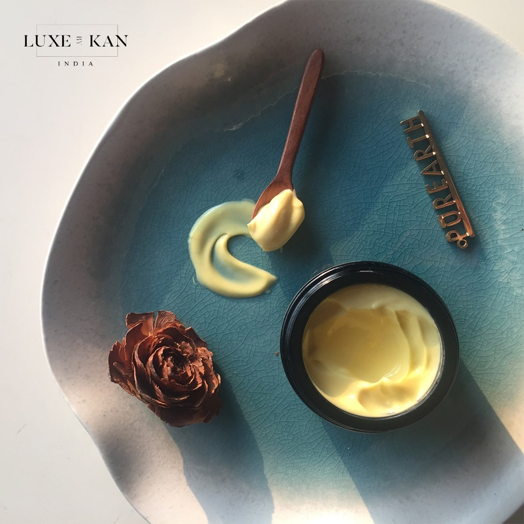 A round, blue ceramic plate holds a jar of creamy moisturizer, a wooden spoon, and a dried rose, with branding "Luxe and Kan."