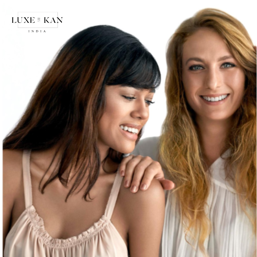 Two women stand closely together, one with dark hair and the other with blonde, against a bright, neutral background.