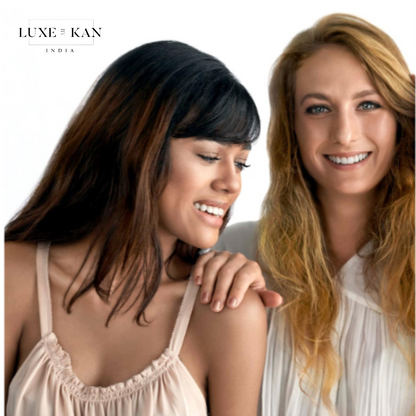 Two women stand closely together, one with dark hair and the other with blonde, against a bright, neutral background.