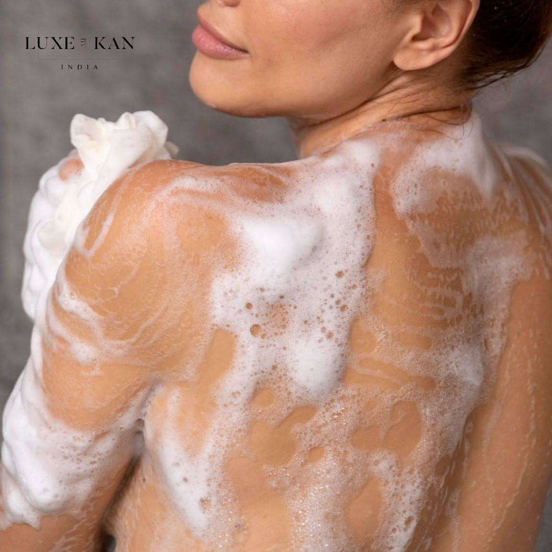 A woman’s shoulder and back covered in soapy bubbles, holding a white loofah, against a soft gray background.