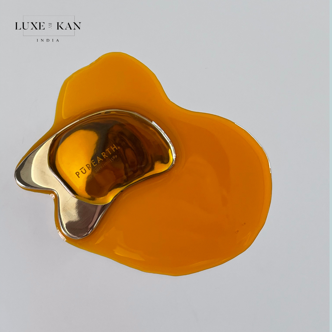 A sleek gold kwansha facial beauty skincare coin rests on a glossy, vibrant orange serum, with branding visible on the tool's surface.