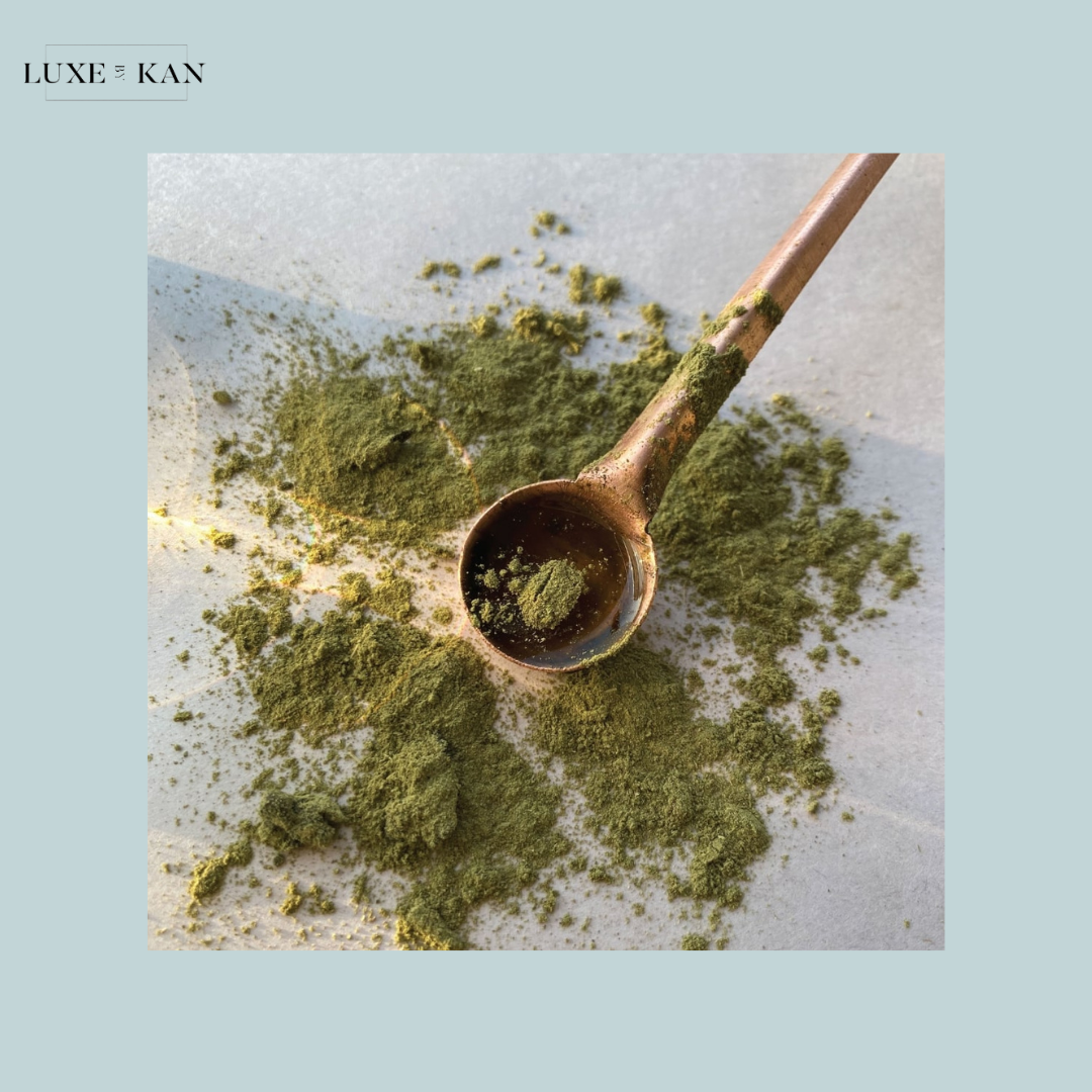 A close-up of a bronze spoon filled with green powder, surrounded by scattered green powder on a light surface.