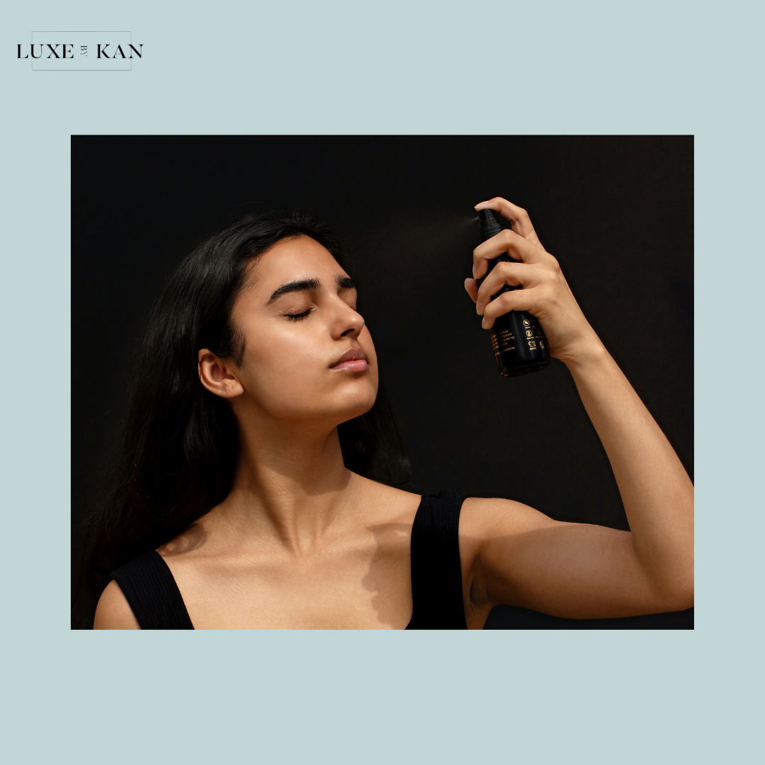 A woman with dark hair is spraying a sambac botanical essence product from a black bottle, wearing a black tank top against a dark background.