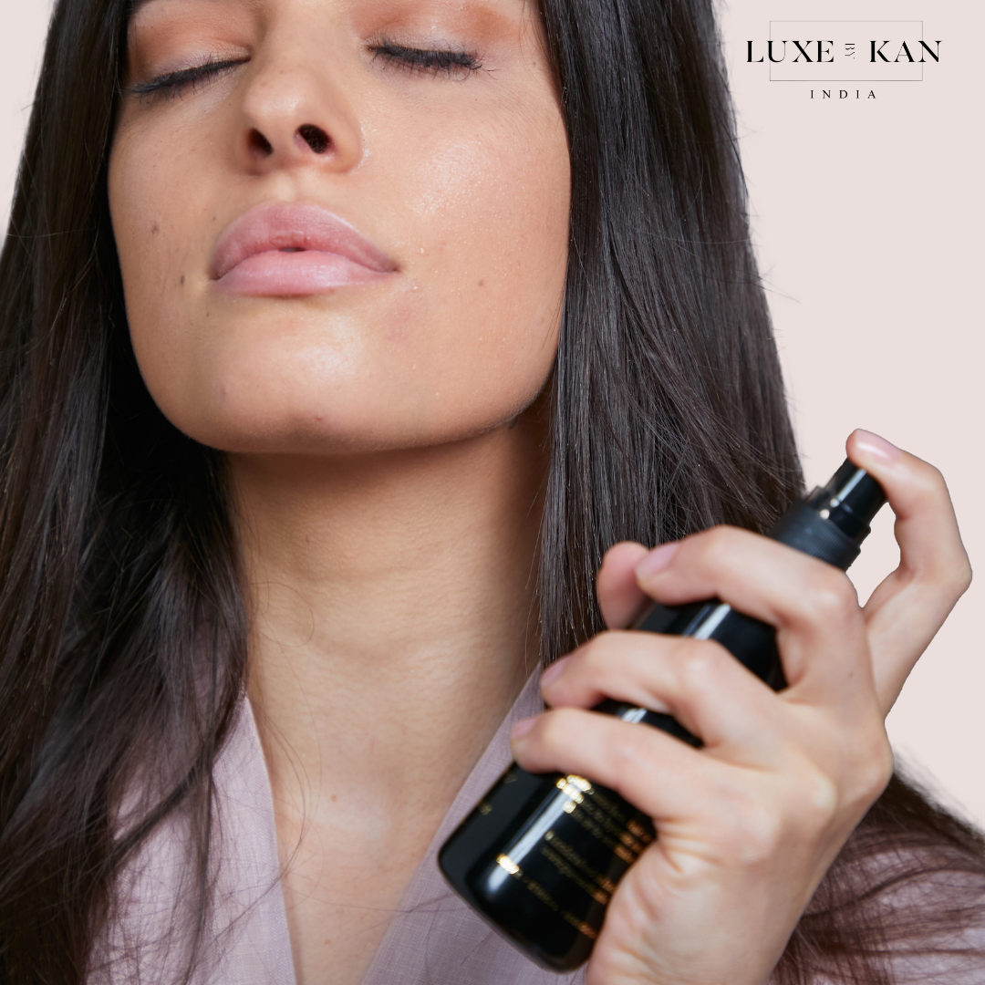 A person holds a sleek black spray bottle, preparing to apply a beauty product, with long, dark hair and a soft-colored robe.