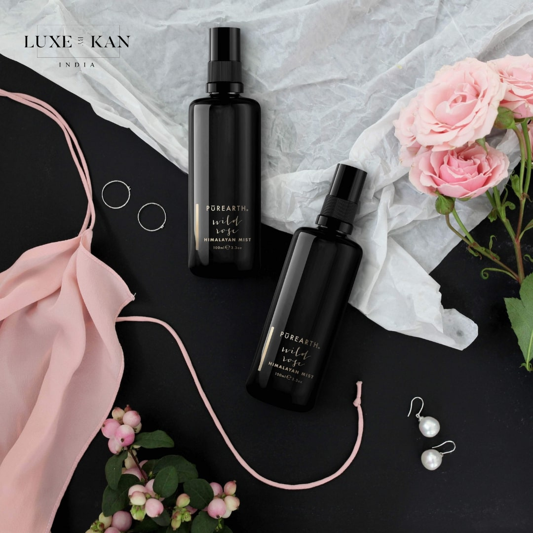 Two black bottles of Purearth Himalayan Mist, surrounded by pink roses, jewelry, and soft fabric, on a dark background.