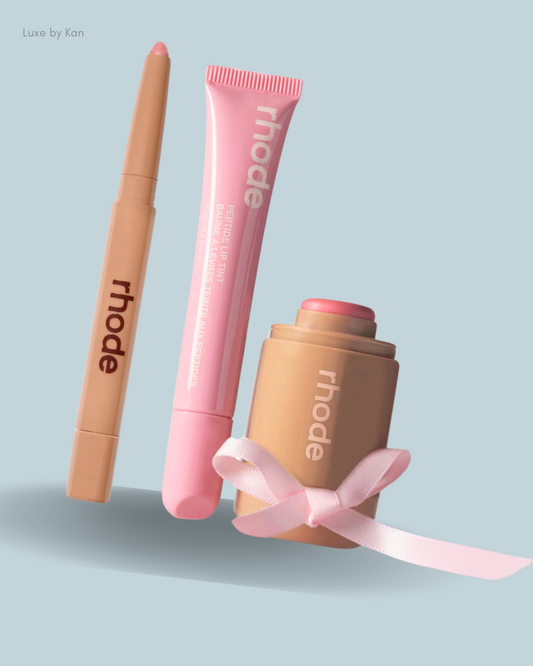 Three rhode beauty products: a lip tint pen, a pink lip balm tube, and a stick blush, elegantly arranged with a pink ribbon.