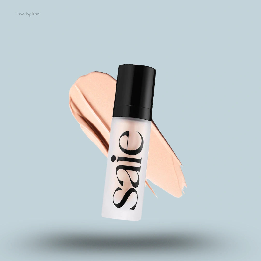 A matte white bottle of SAIE Glowy Super Gel in a frosted tube with a black cap, floating on a peach foundation swatch.