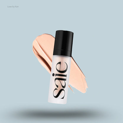 A matte white bottle of SAIE Glowy Super Gel in a frosted tube with a black cap, floating on a peach foundation swatch.