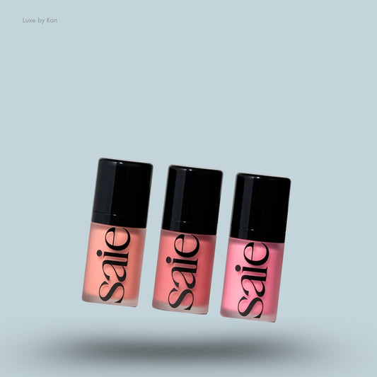 Three bottles of SAIE Mini Dew Blush Trio in shades of peach, coral, and pink with black caps on a blue background.