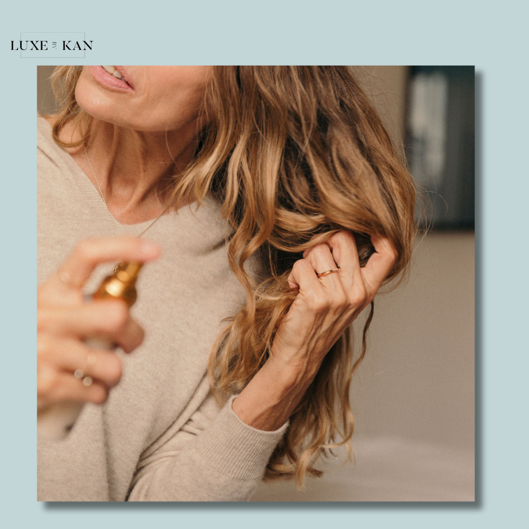 A woman in a beige sweater sprays a hair product while gently holding her wavy, golden hair, showcasing a healthy haircare routine.