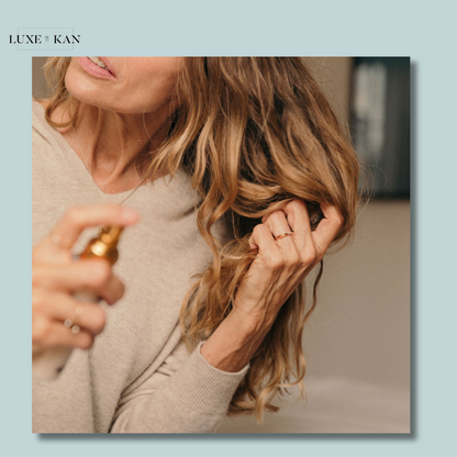 A woman in a beige sweater sprays a hair product while gently holding her wavy, golden hair, showcasing a healthy haircare routine.
