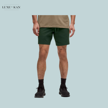 A man stands wearing green shorts and a beige shirt, paired with black socks and shoes.