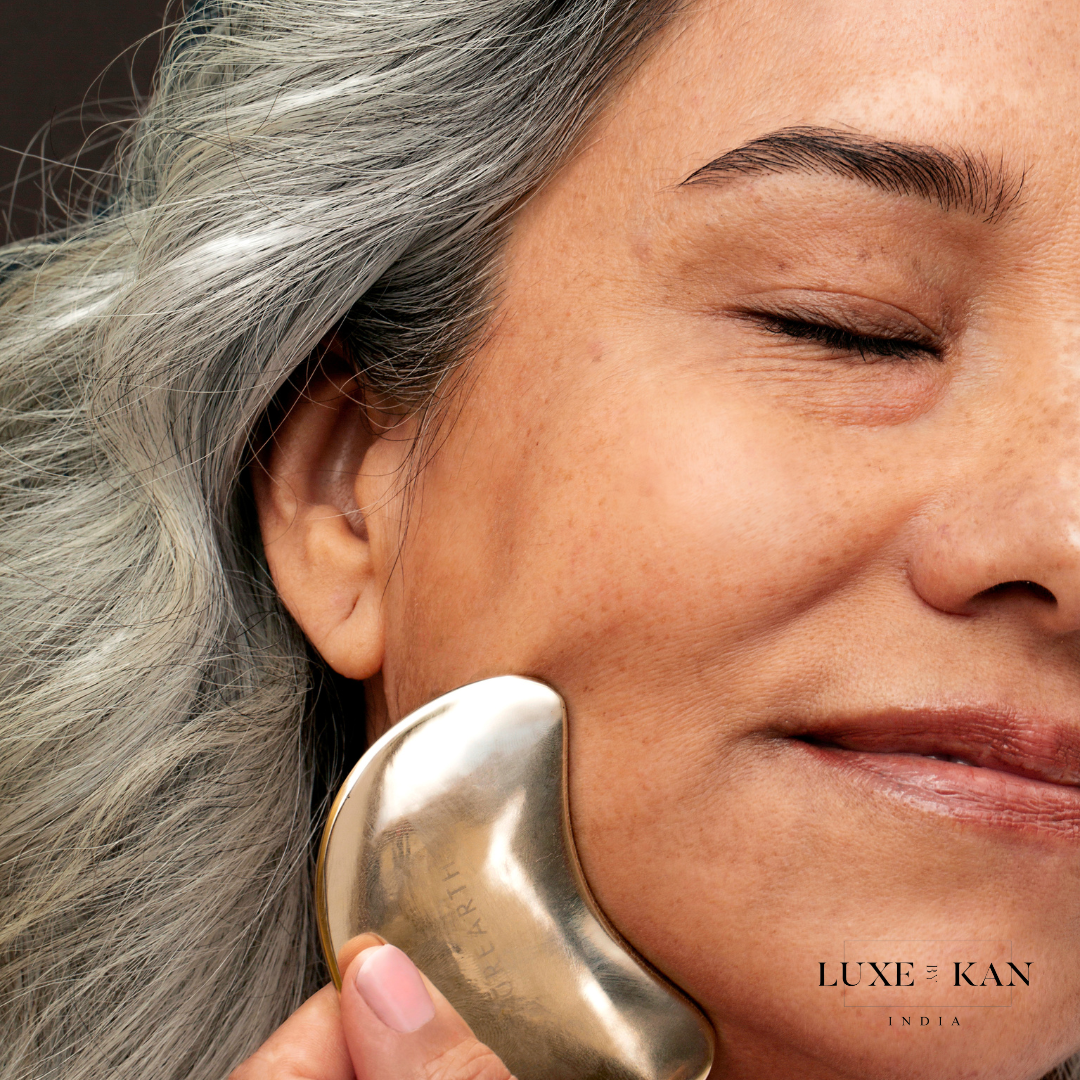 A woman with gray hair uses a Purearth kwansha facial beauty coin on her cheek, eyes closed, with the Luxe Kan India logo in the corner.