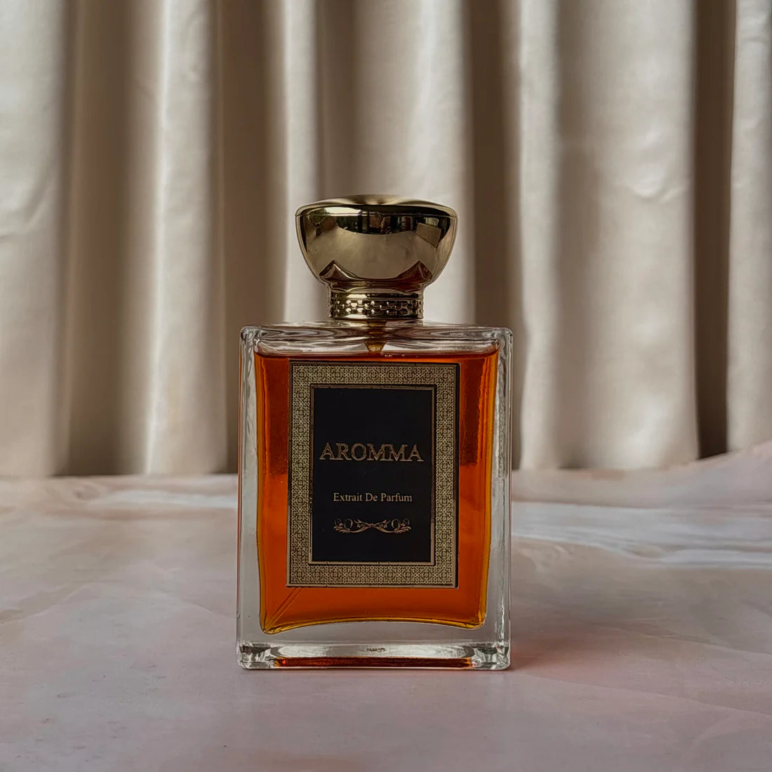 Aromma White Amber Extrait De Parfum in a clear glass bottle with a substantial gold cap against a cream-colored draped fabric backdrop.